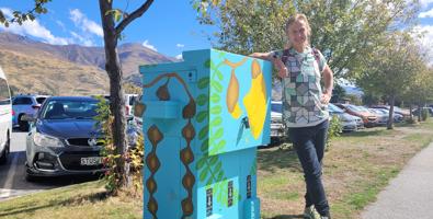 Streetart: Wānaka artist, Chrissy Wickes standing next to a switchbox painted with kowhai at various stages of flowering and seeding