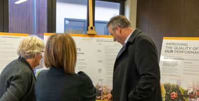 Photo of from a community event. Three people with their backs to the camera are looking at an information board.