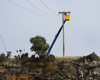 Photo shows a worker in a cherry picker basket at the top of a power pole which is at the top of a rugged cliff.