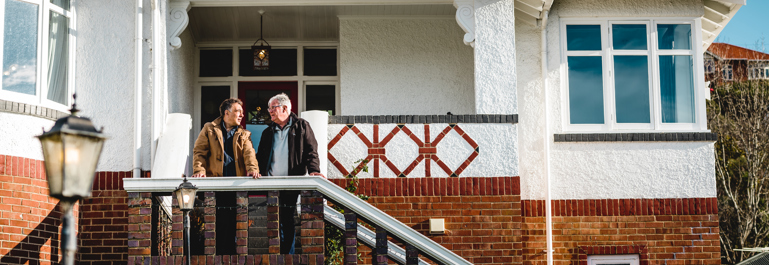 Photo of two men standing on the steps of a brick house leaning on the handrail talking to each other.