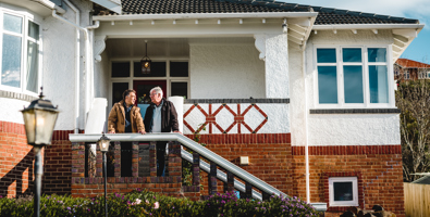 Photo of two men standing on the steps of a brick house leaning on the handrail talking to each other.