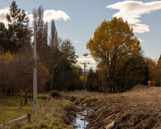 Photo shows a helicopter in the background with power lines in between trees on either side of a creek.