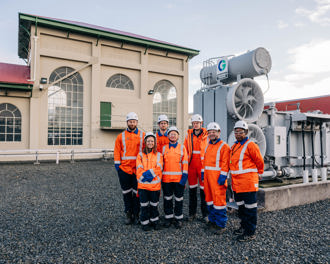 Group of people wearning orange personal protective high vis clothing and hard hats standing in front of a substation