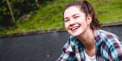 Photo of a young woman outside, crouching down, looking up at something behind the camera, smiling. 