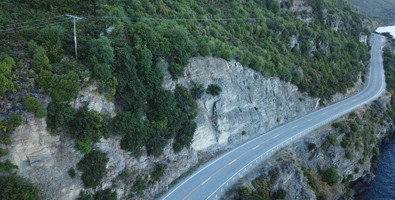 Photo of a long stretch of road halfway up the side of a hill, above a body of water. Above the road is sheer cliff face , trees and then a power line.