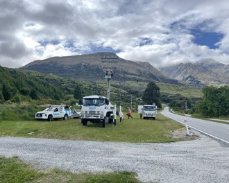 Photo shows two Delta trucks and a ute on the side of the state highway with two workers near the top of a power pole working