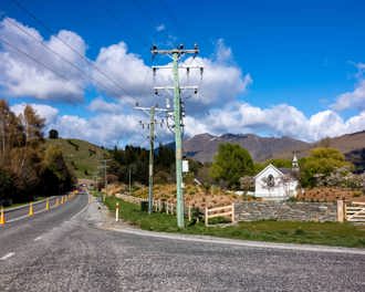 Photo shows a long stretch of road with road cones along the centre line and new power poles on the side of the road. Mountains in the background and a small church in the midground.