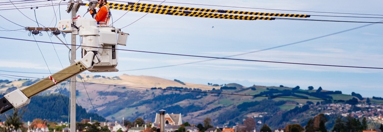 A person in a cherry picker at the top of a power pole working, with city and rural-scape behind them.