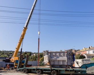 Image shows a crane lifting a large transformer off the back of a truck at construction site of Andy Bay substation upgrade.