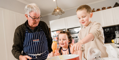 Photo of a grandfather wearing an apron, standing with two young children in the kitchen. They are baking