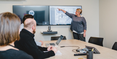 Meeting room, woman at the front of a team of people pointing to plans on a large screen